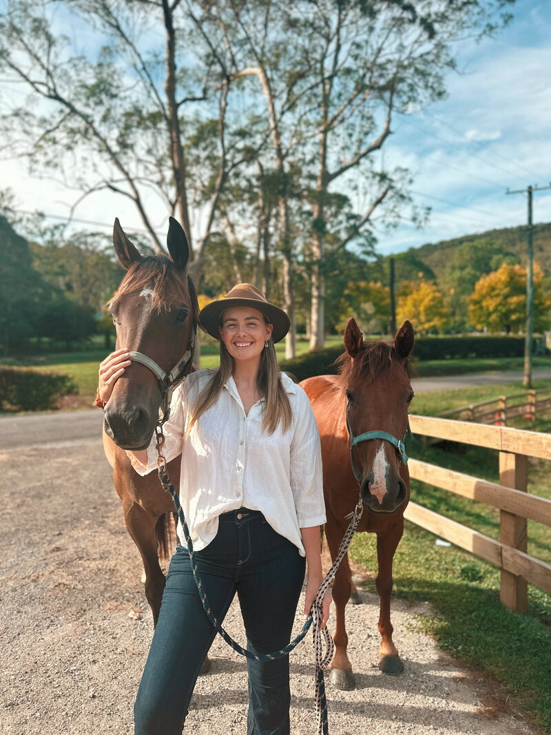 A young boy in a blue top, black pants and a cargo print hat brushing a dark brown horse