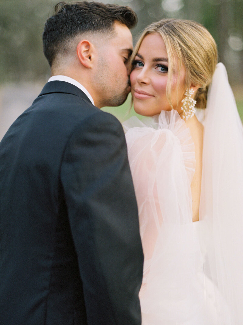 Groom kisses his bride's cheek as she looks over her shoulder