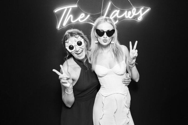 bride and friend posing against a black backdrop with a custom neon sign on, during a Hyde House wedding event 