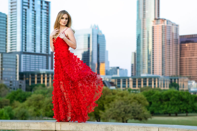 Senior girl with red dress posing on a ledge in front of a city scape
