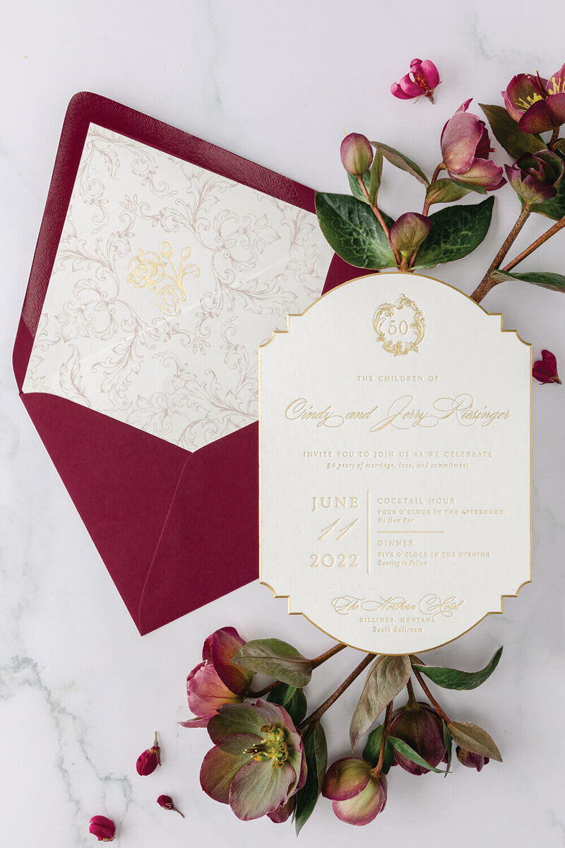 Luxury gold foil invitation with custom monogram, scroll details, and burgundy envelope liner designed by Seventh and Anderson for a 50th wedding anniversary celebration.