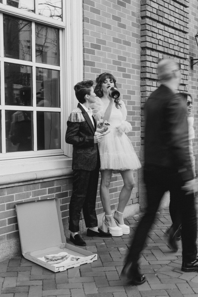 Black and white image of nonbinary newlywed couple celebrating their marriage leaning against a building in Georgetown DC eating pizza and sipping champagne from the bottle while blurred people pass by