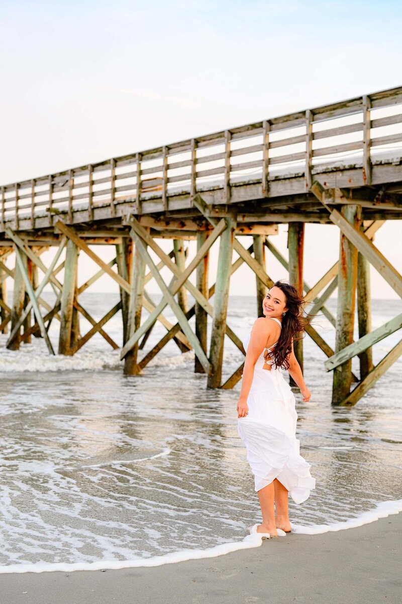 Waxhaw senior plays on the beach next to a charleston, sc pier during her graduation session wearing a white dress.