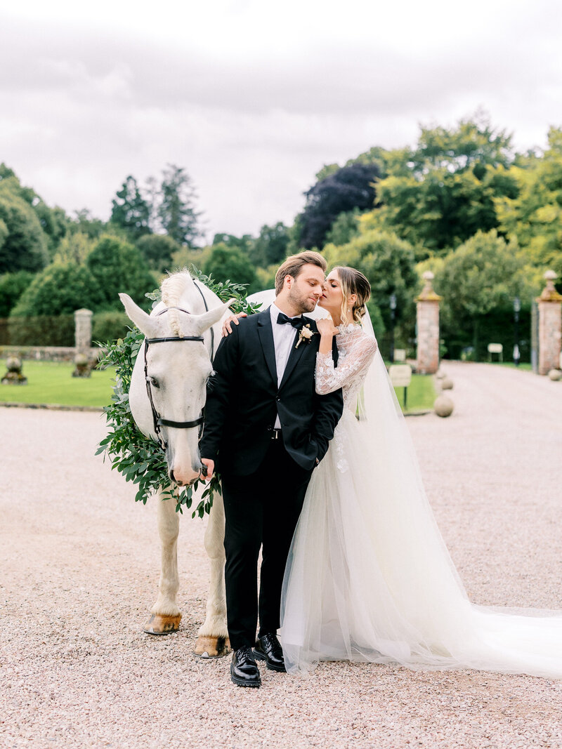 Bride kisses groom on the cheek while groom holds onto the reins of a while horse with greenery wreath around its neck