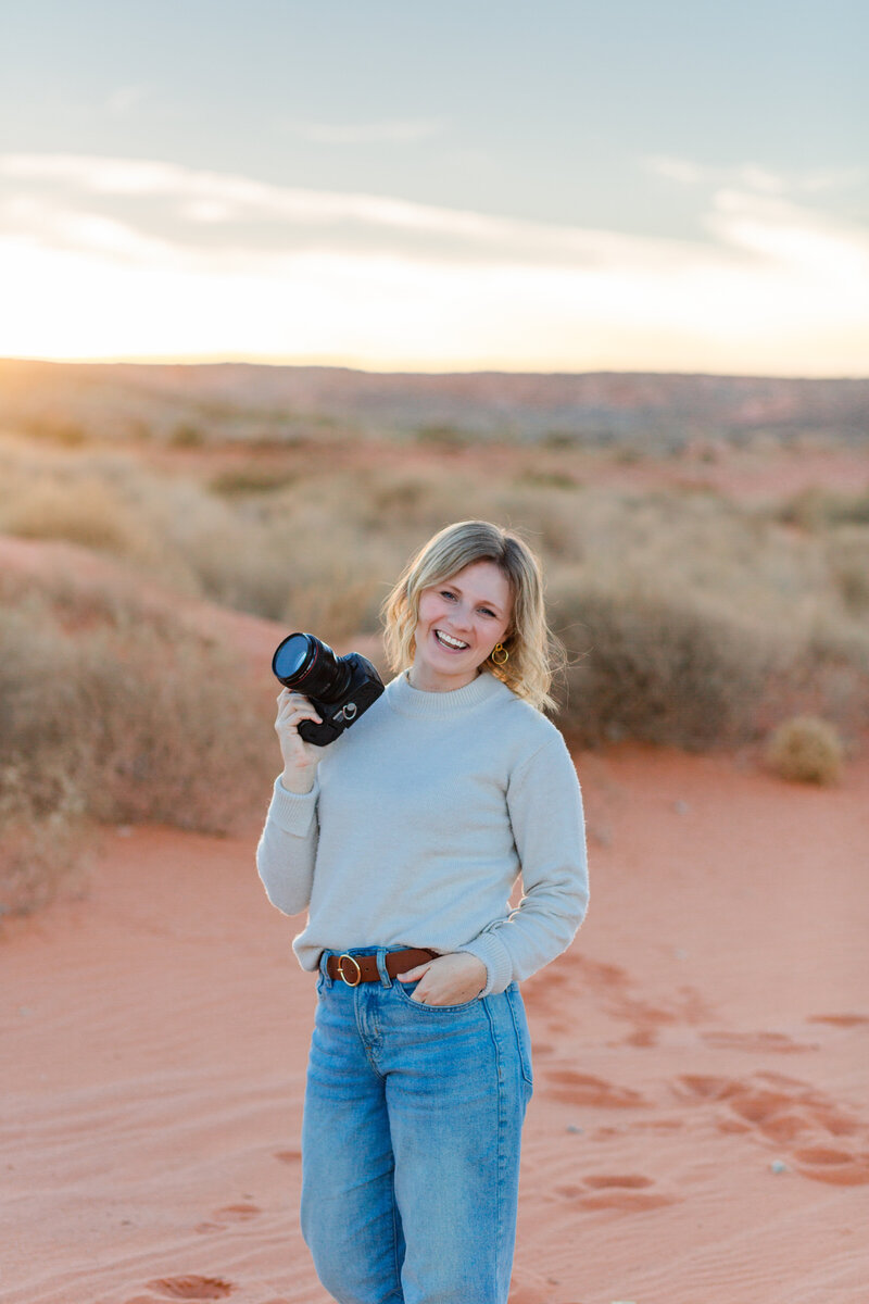 St. George photographer girl standing in the sand at sand hollow state park holding the camera and smiling