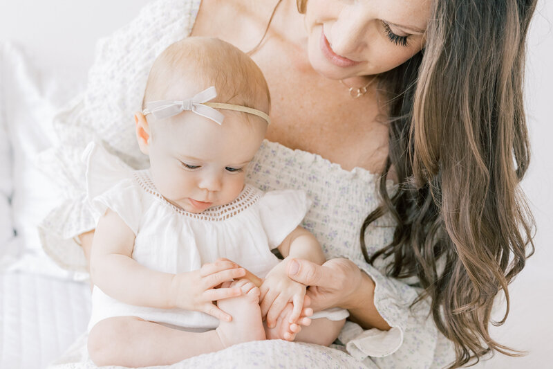 A mother and her infant daughter sit on a bed playing with the infant toes during a session with a Charlotte Family Photographer