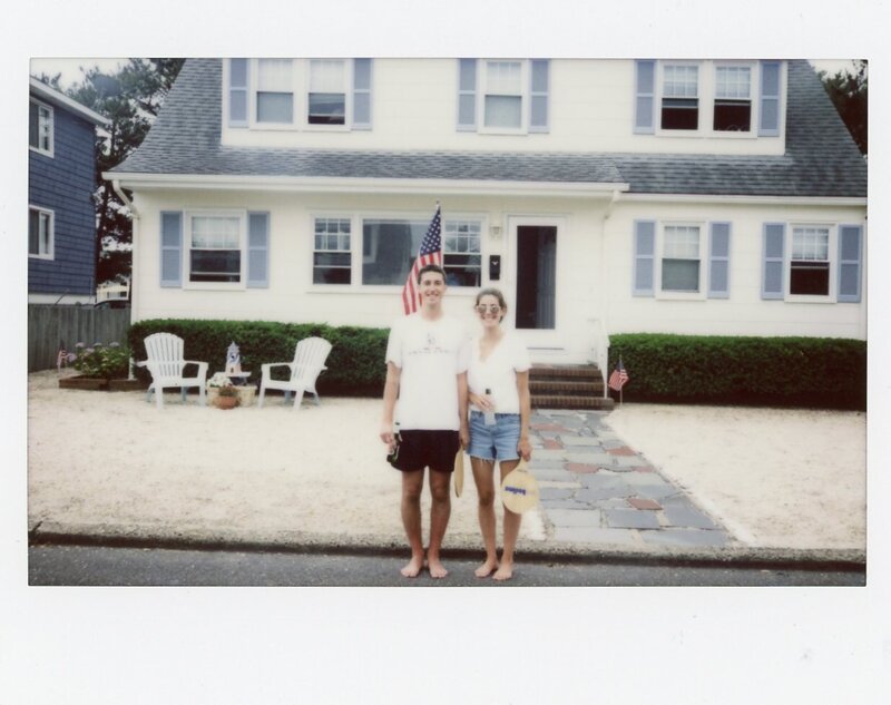 Polaroid of young couple smiling and standing in front of white California beach house with blue shutters while holding paddles