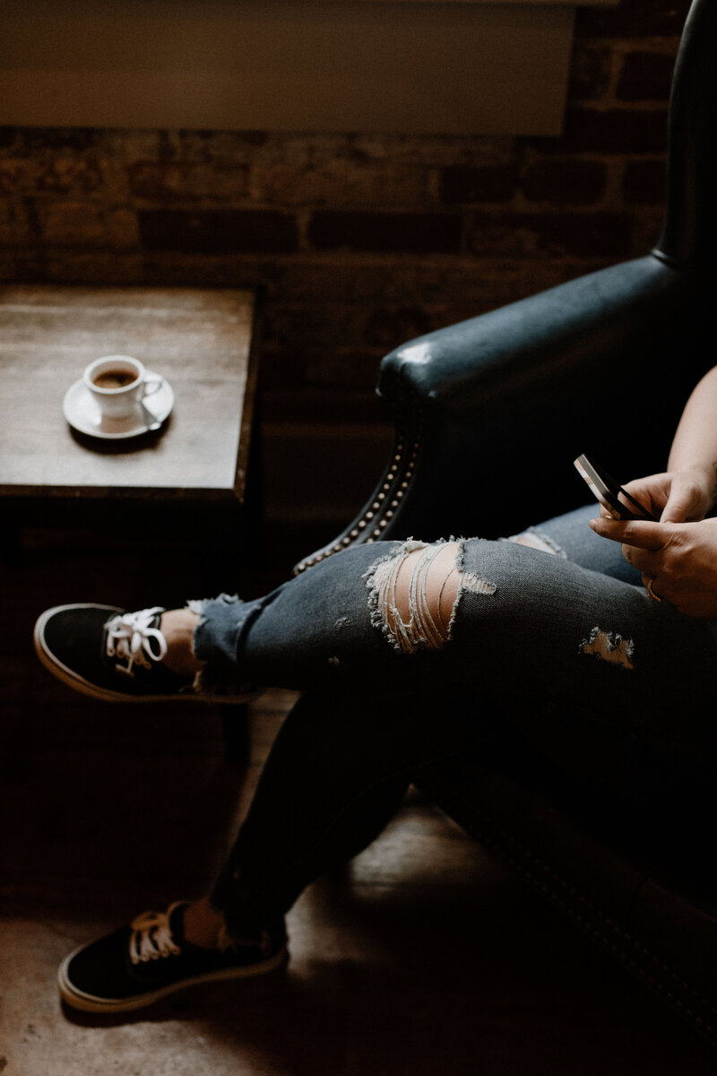 A person wearing ripped jeans and black sneakers sits in a dark leather chair, holding a phone—perhaps browsing a white label design course. A small cup of coffee rests on a wooden table nearby in the cozy, dimly lit room.