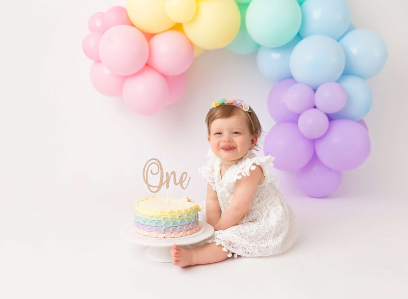 Baby girl at her cake smash session with Rochel Konik photography wearing white dress and sticking  her tongue out  rainboy balloon garland theme