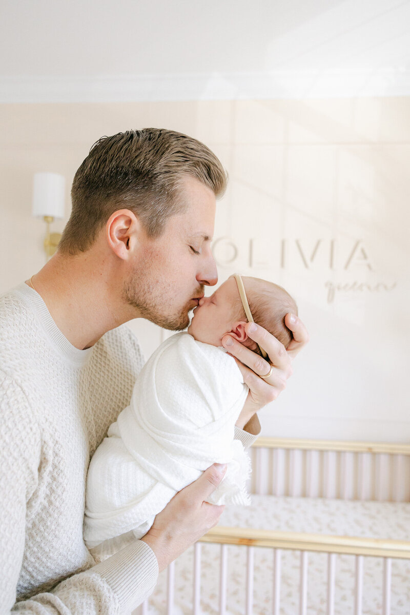 A father gently kisses his swaddled newborn daughter, Olivia, in her soft-toned nursery during an in-home newborn photography session in Fairfield County, CT. Captured by Nicole Neira Photography, this image celebrates early fatherhood and heartfelt connection.