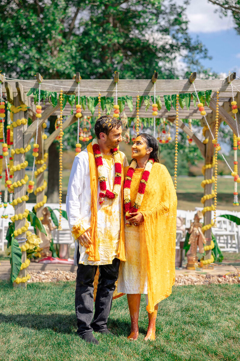 A couple stands smiling under a decorated wooden canopy. They are wearing yellow and red garlands and traditional attire. The background features greenery and hanging decorations, with chairs arranged nearby.