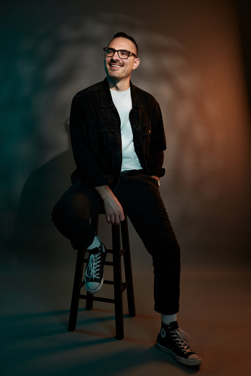 Stylish full-body studio portrait of Mike Moretti seated on a stool, wearing a dark jacket and trousers, with dynamic gradient lighting, captured by Ylva Erevall.