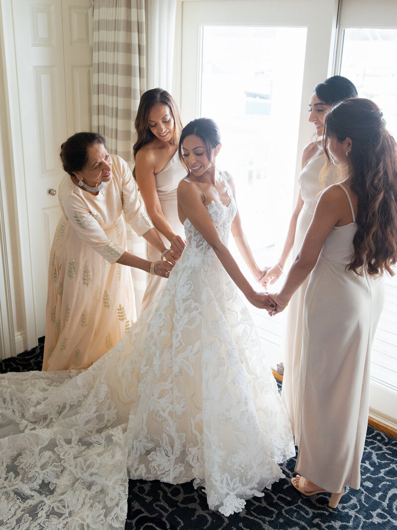 Bride in a lace strapless gown holds hands with her sisters while her mother zips up her wedding gown.