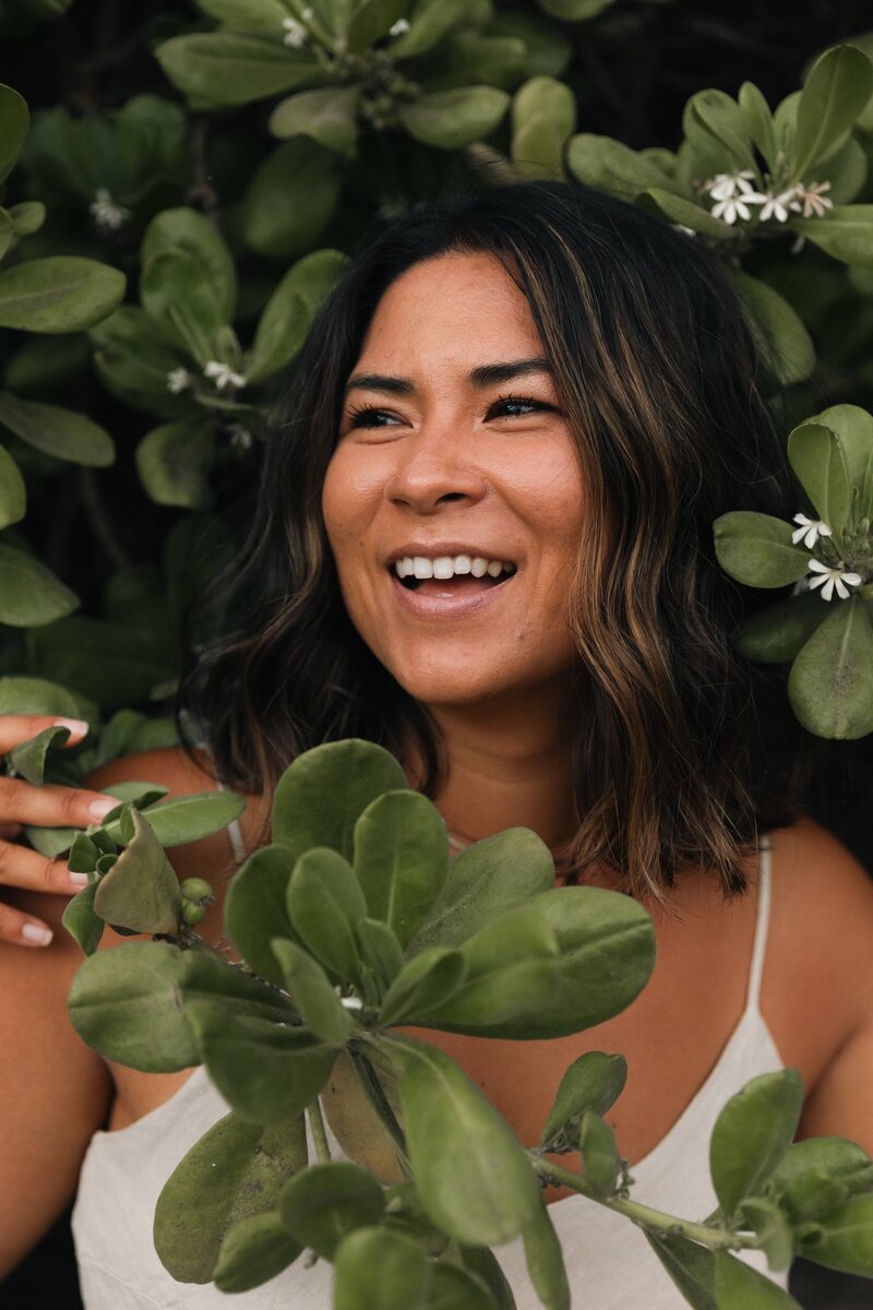 Amber, surrounded by tropical greenery, smiles while looking off camera to the left mid laugh.