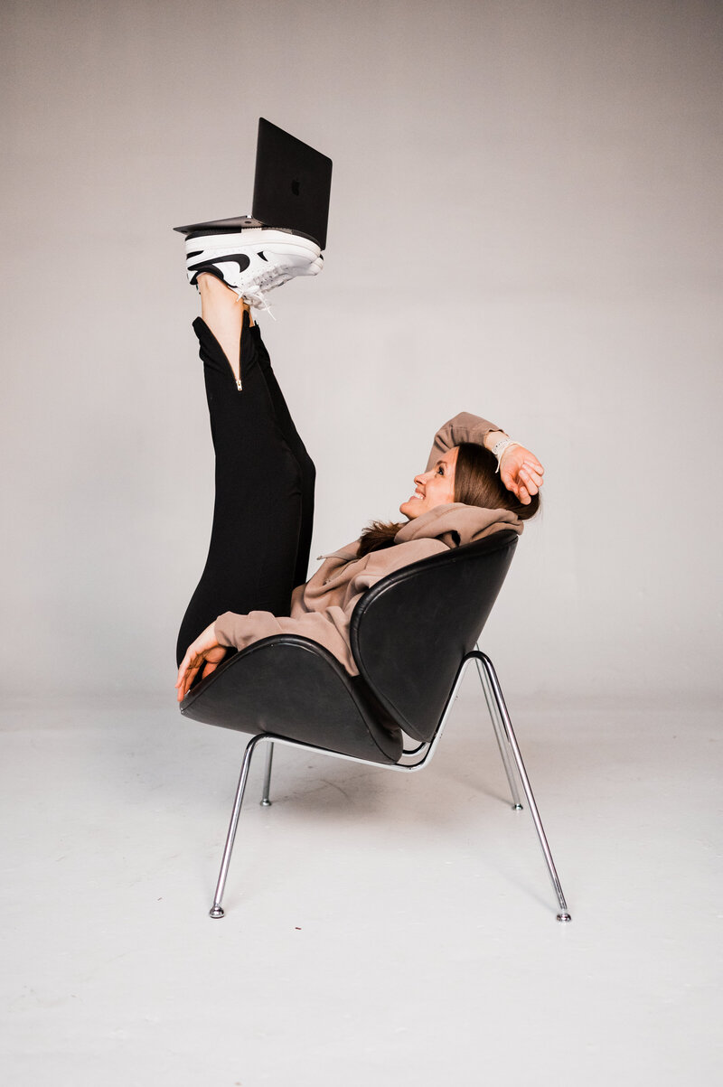 Woman smiling in a chair balancing a laptop on her feet, participating in a photography mentorship program that helps her run a business with more free time.