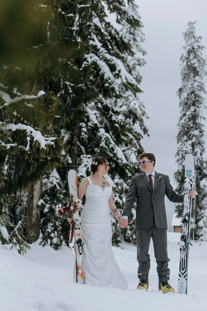 Bride and groom stand in snow in winter holding skis wearing heart shaped sunglasses