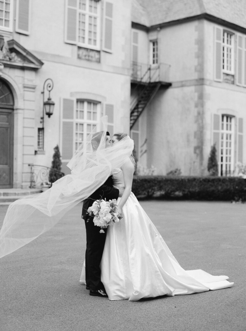Rhode Island Wedding Photographer | A bride and groom share a kiss outside a historic building. The bride's veil flows in the wind, creating a sense of movement and romance. Black and white photo.