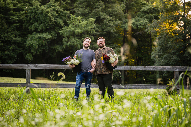 A gay couple stand in a field holding a bucket of flowers for their anniversary adventure portrait session at their farm in Roanoke, Virginia.