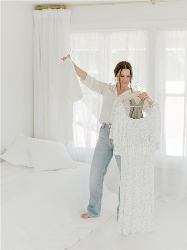 Photographer holds up two floral dresses in a bright, white studio while planning outfits for a photo session in Kansas City.