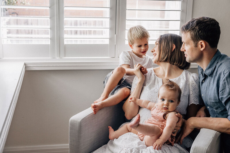 Family gather and sit together on couch in front of window at home photography family photography session