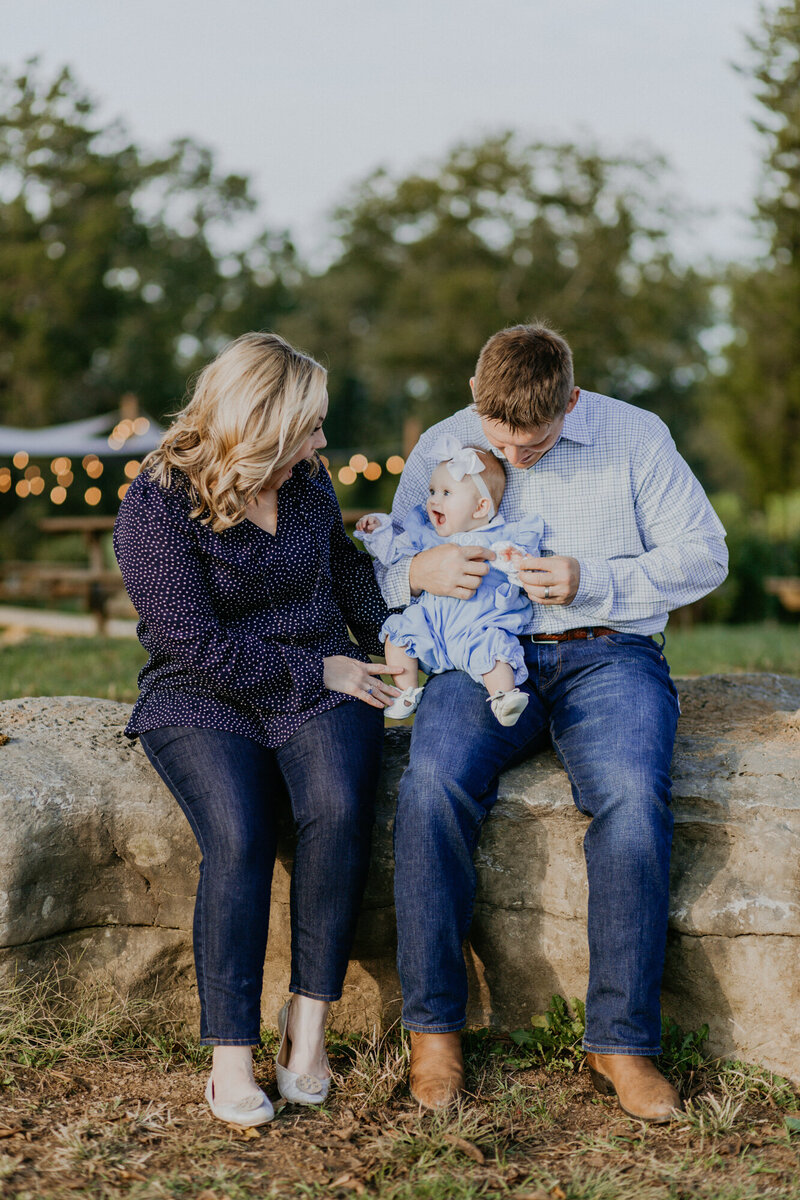 A mother and father hold their little baby girl on their knee while sitting on a stone fence