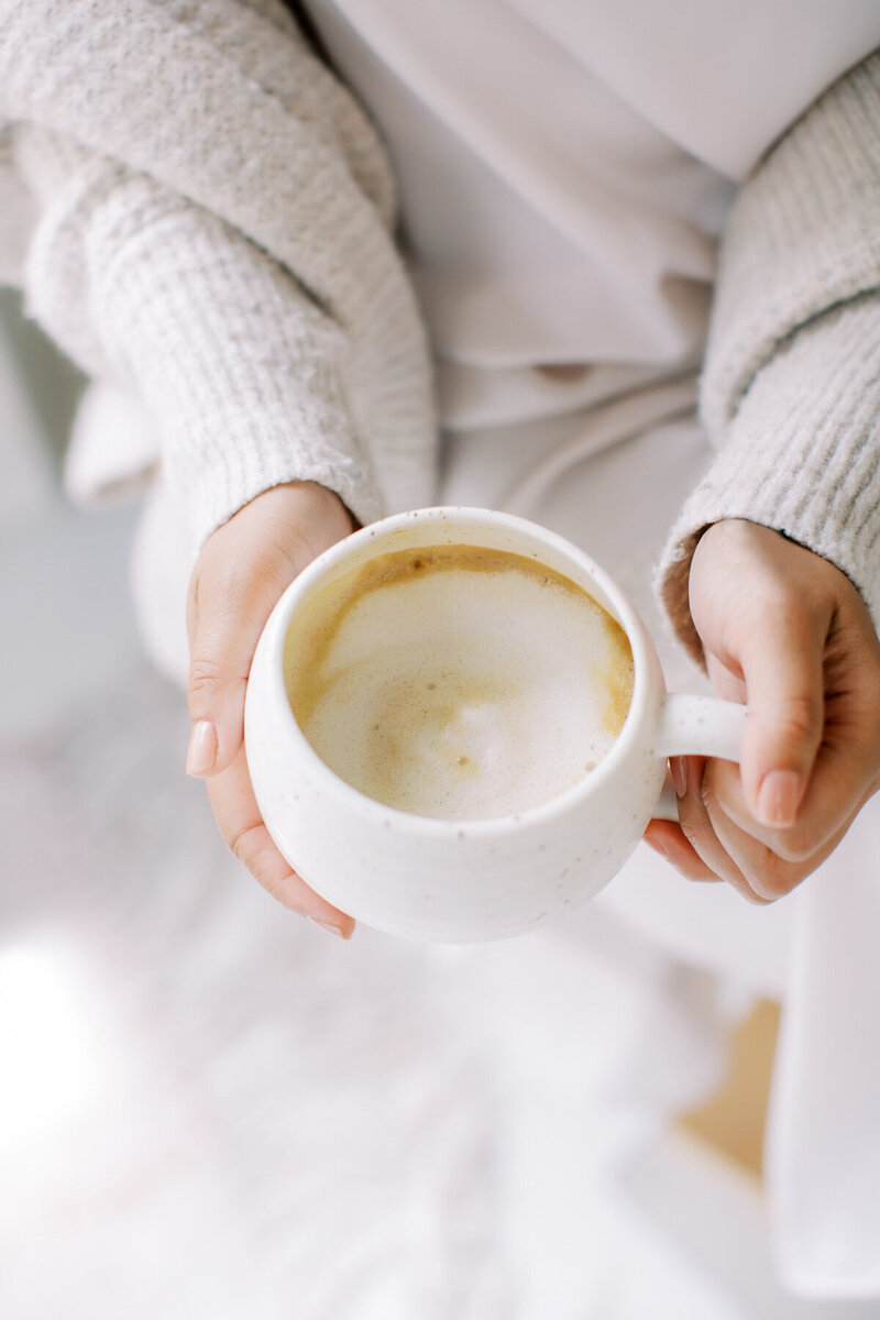 A person holding a white ceramic cup filled with frothy coffee. The individual's hands are wrapped around the cup, and they are wearing a cozy, light-colored sweater. The background is softly blurred, suggesting a warm and relaxed atmosphere.