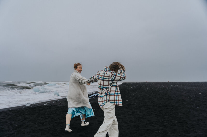 Two lesbians walk down the black sand beaches of Iceland while smiling 