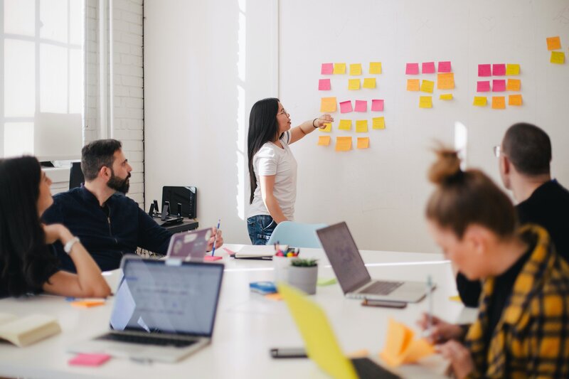 une femme debout lisant des post-it devant 4 adultes sur un bureau