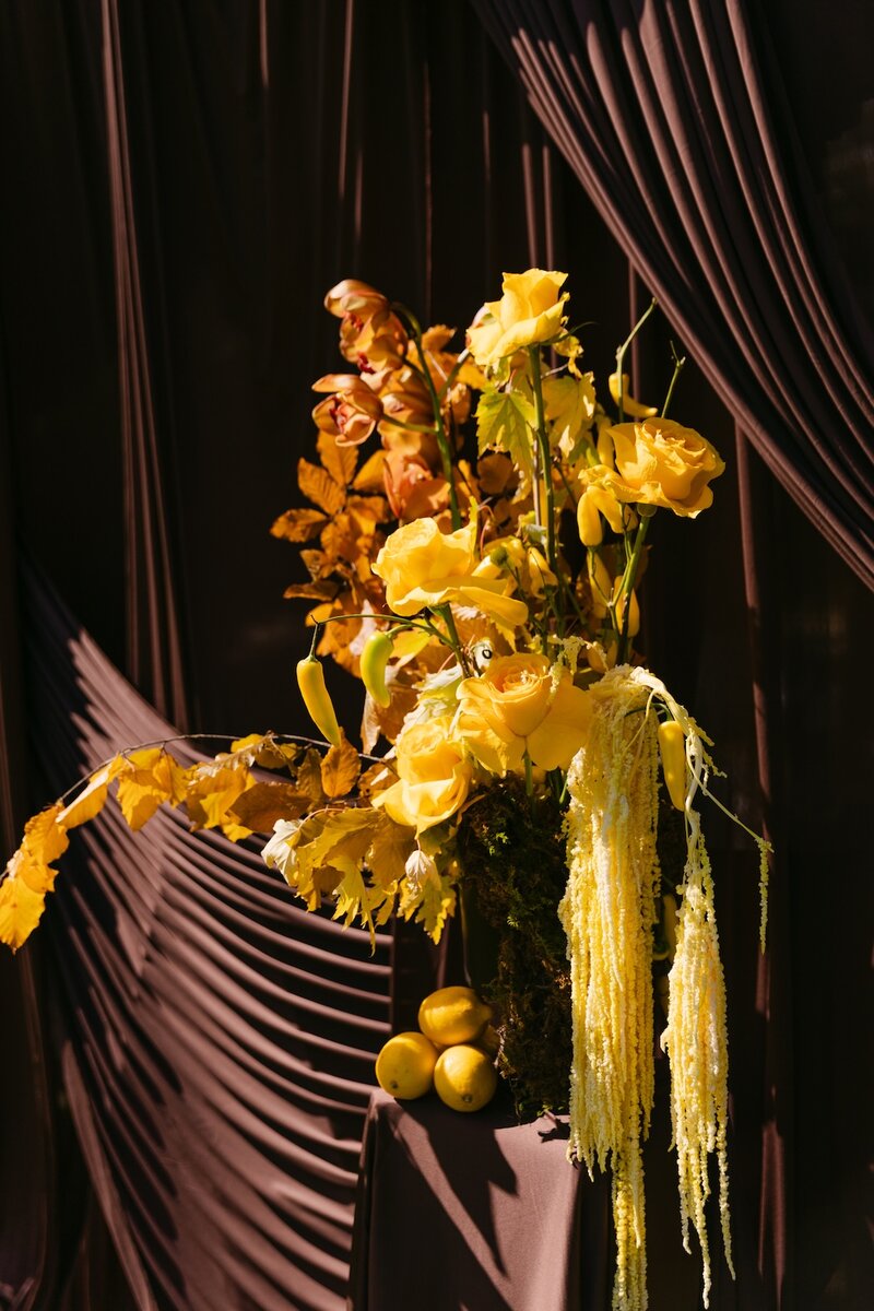 Colorful yellow wedding florals designed creatively with foods against a chocolate brown drape for a luxury wedding ceremony at Oceana's Gardens in Boulder, Colorado.