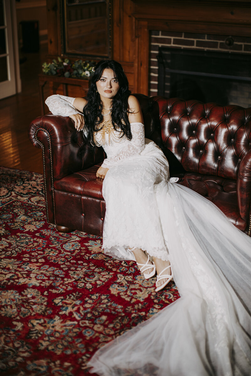 Bride in long white gown sitting gracefully on a vintage red leather sofa in a warm, classic room