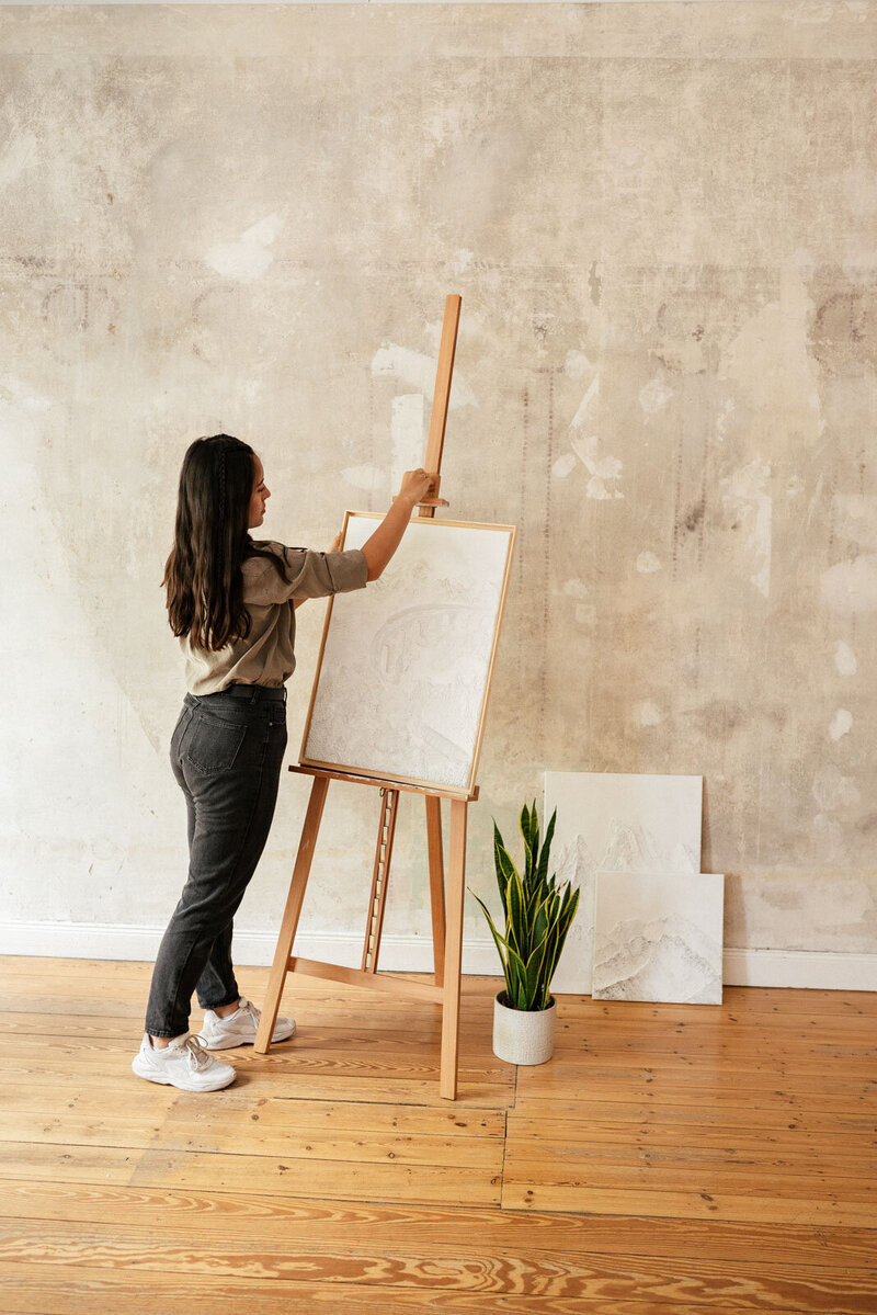eine Künstlerin steht im Atelier und positioniert ihr Kunstwerk auf einer Staffelei