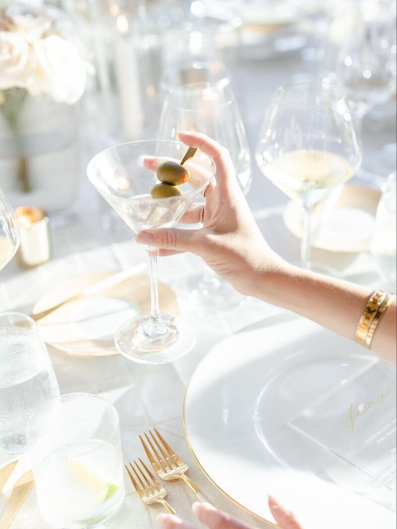 A wedding guest holds a martini glass amidst white and gold dinnerware