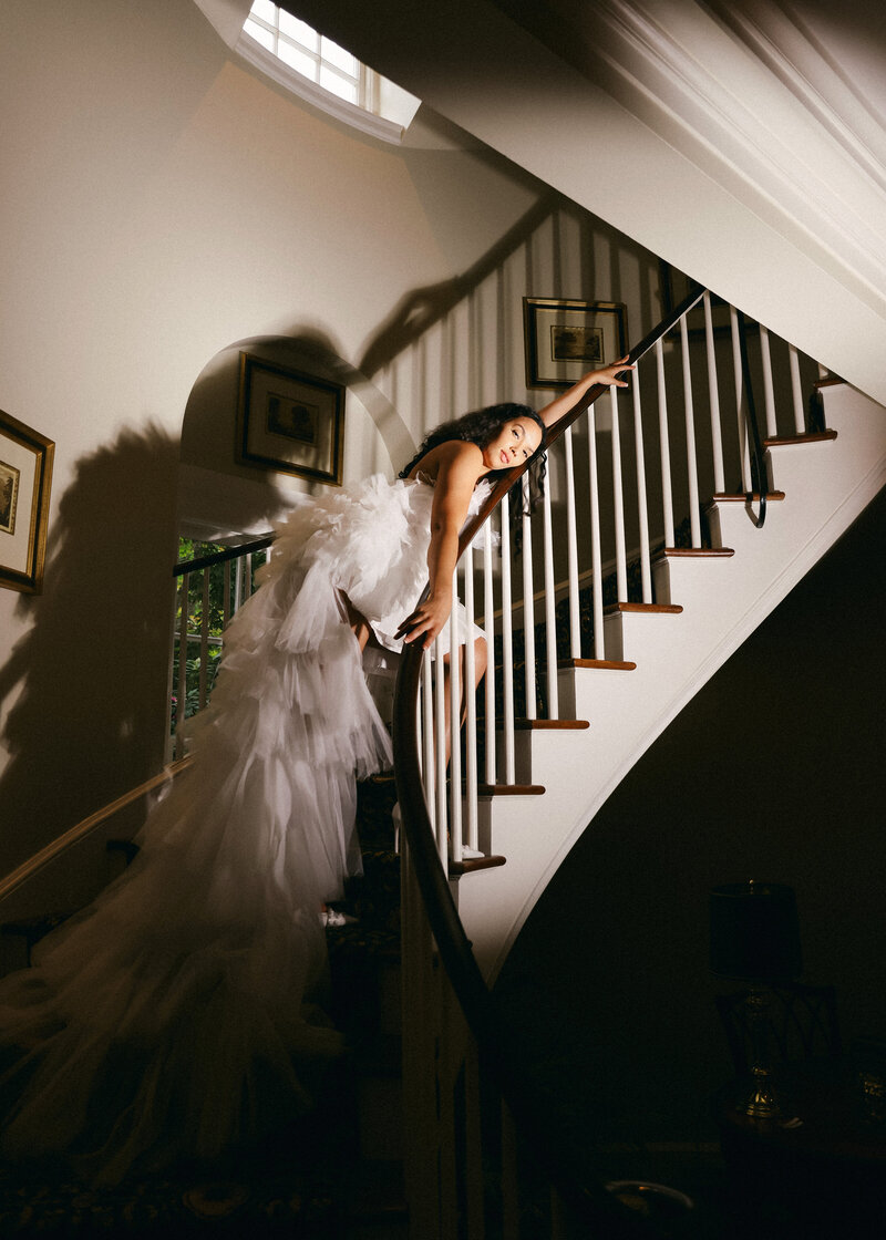 Brittany gracefully walks down the stairs at Boxwood Estate, preparing for her wedding day. Her hair is pulled back into an elegant bun, and she wears a delicate white lace robe. The soft, natural light illuminates the moment, highlighting her serene expression as she makes her way toward the ceremony. The elegant interior of the estate provides a beautiful backdrop, adding to the sense of anticipation and excitement.