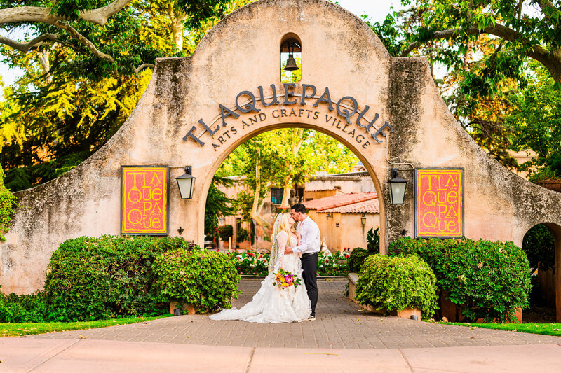 Wide shot of bride and Groom in wedding attire kissing under Tlaquepaque Arts and Craft Village arch walkway in Sedona with green trees and bushes