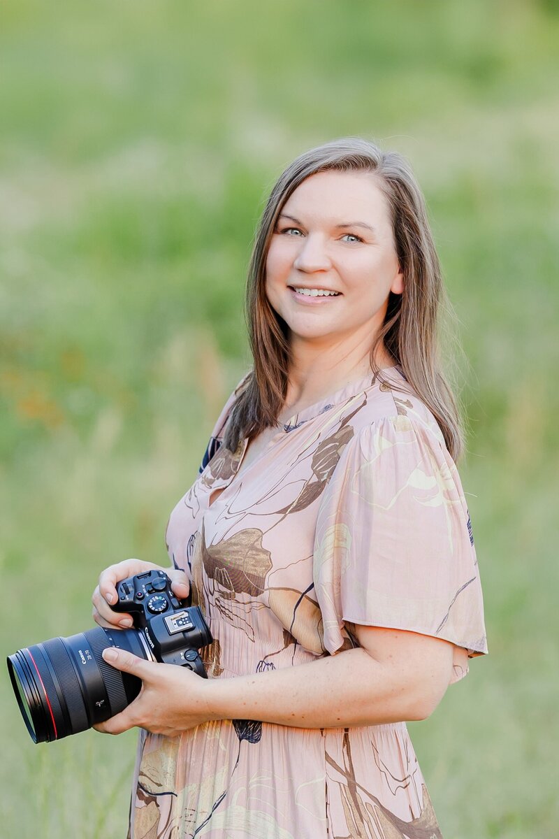 A women in a dress holds a camera while standing in a field of flowers for a portrait.