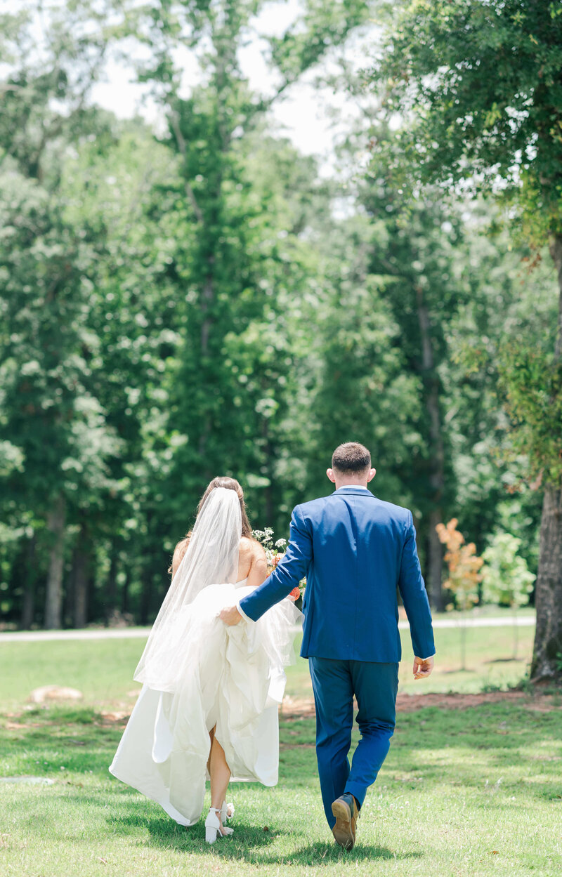Bride and groom walking away from camera while groom  holds brides dress