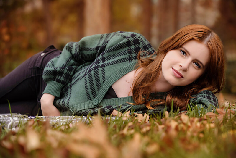 Beautiful HS senior portrait in green cardigan laying on the ground surrounded by golden fall leaves in Ballantyne.