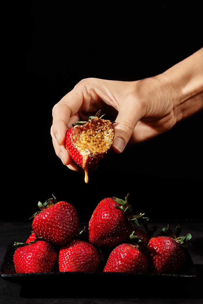 A dramatic close-up of a hand holding a ripe strawberry, dripping with honey. Photographed by Mica McCook, this image highlights the natural sweetness and rich texture of the fruit, creating a visually striking and appetizing scene.