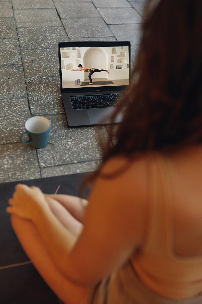 Over-the-shoulder view of a person seated cross-legged on the floor, watching Sarah demonstrating a pose on a laptop, with a serene and focused atmosphere suggested by the clear workspace and a cup of tea nearby.
