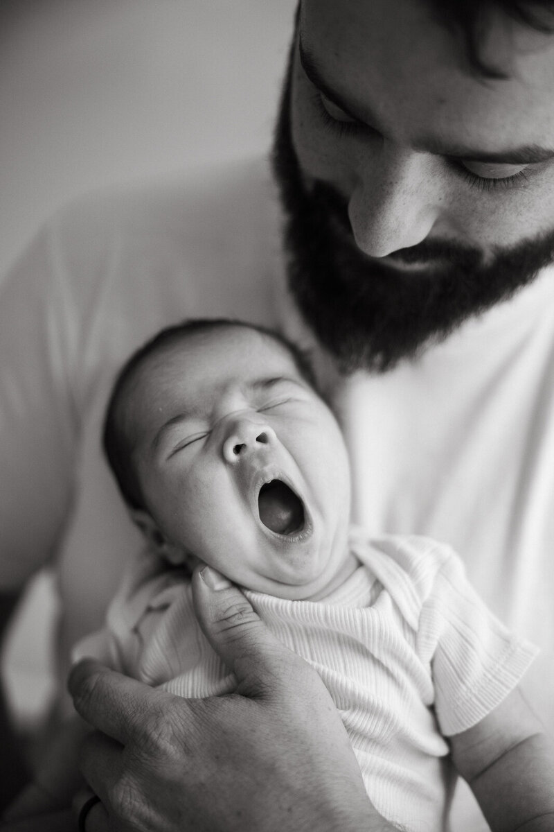 A black and white portrait of a dad holding a newborn who is yawning.