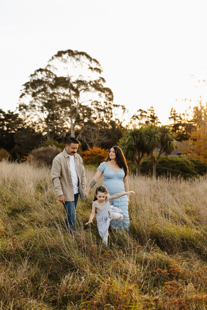 A family of three walking through a field of long grass during a maternity photoshoot with christchurch photographer lucy jane photography