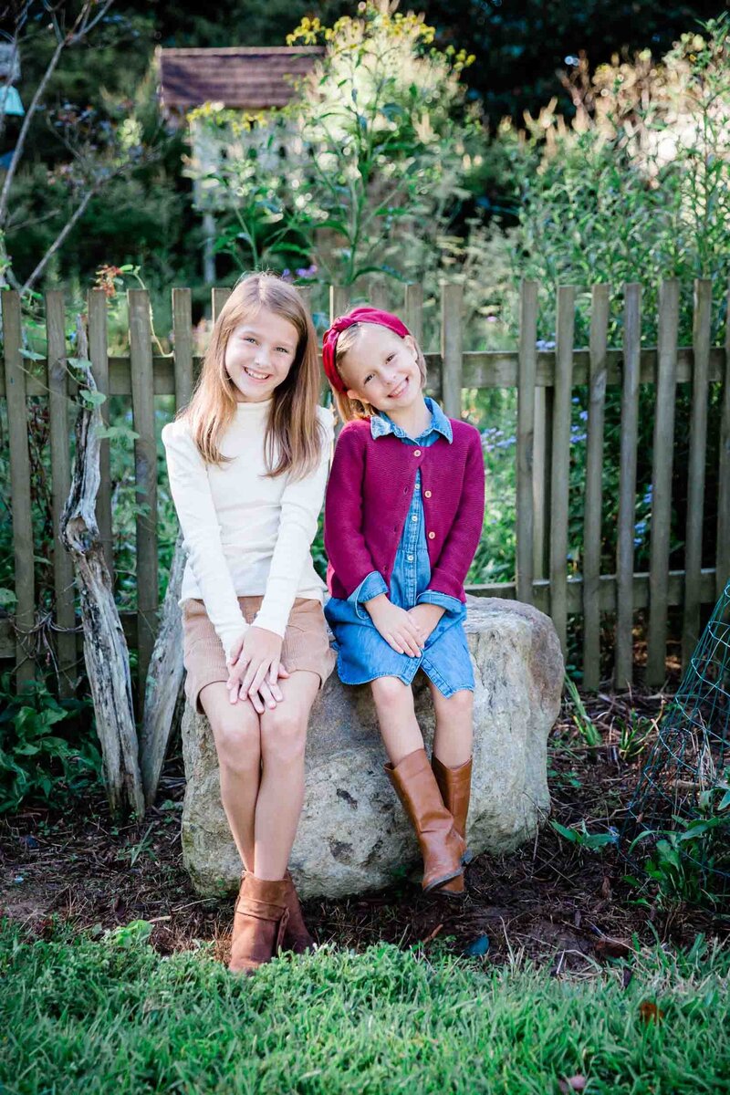 Two sweet sisters sitting on a rock smiling sweetly at the camera during their family portraits.