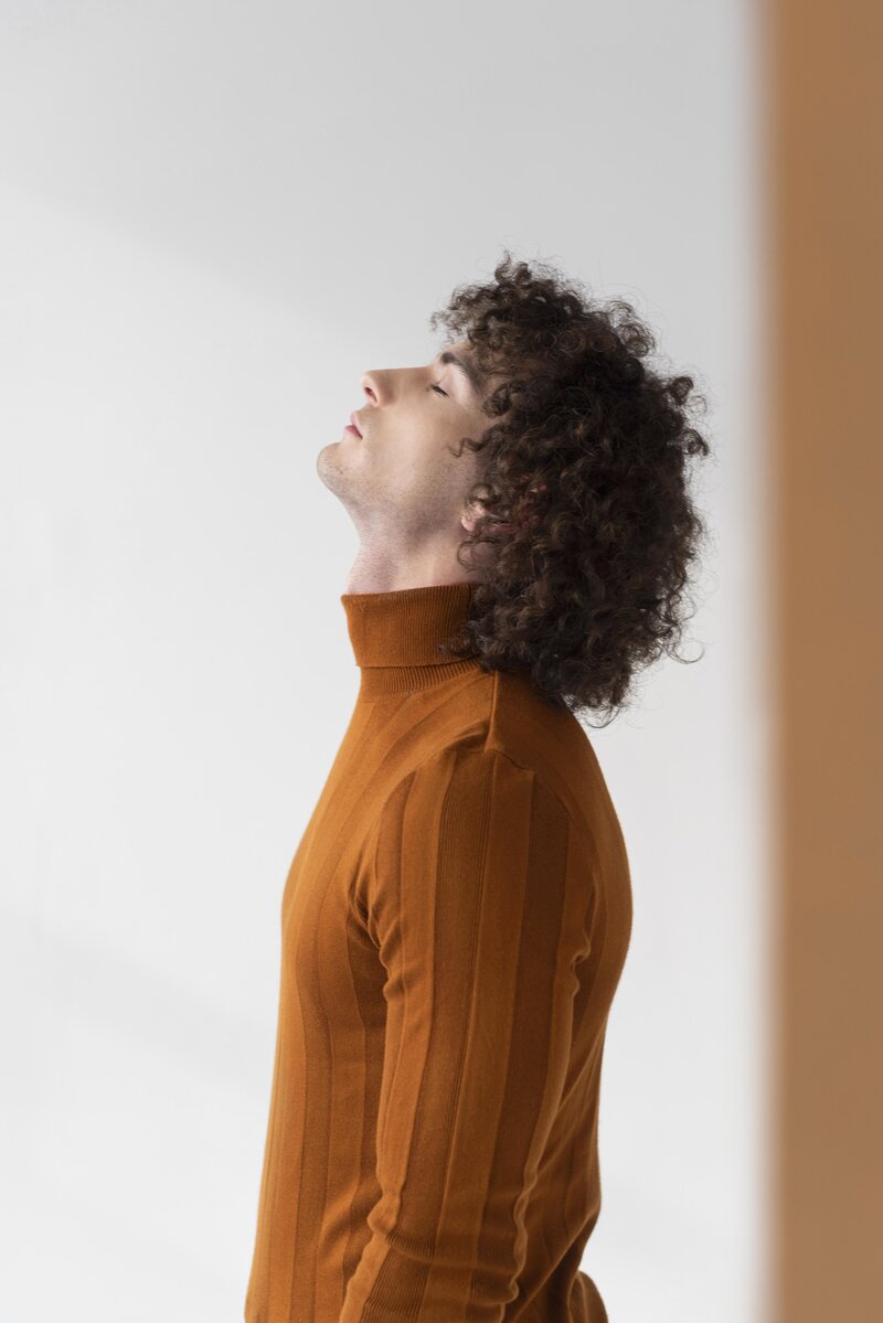 Young man with curly hair and brown top gazing upward in a neutral studio, representing self-reflection and somatic awareness in NYC therapy.