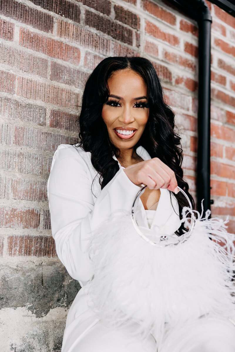 Tiara wearing a white top and pants holding a white feather purse in front of a brick wall