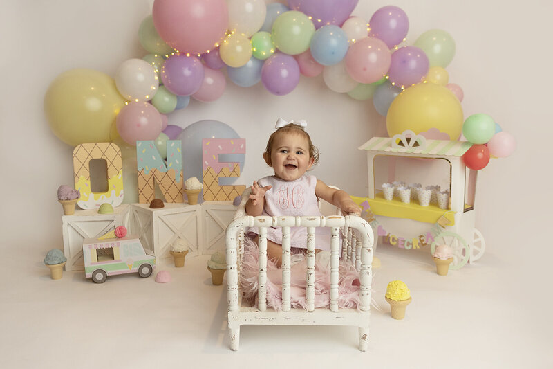A young toddler girl sits in a white wooden crib in a birthday decorated studio
