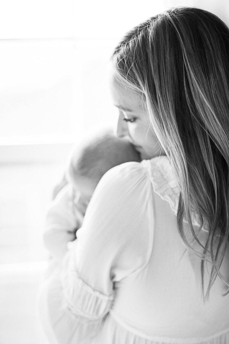 A black and white photo of a mother gently holding her newborn against her shoulder in soft window light during a Kansas City lifestyle session.