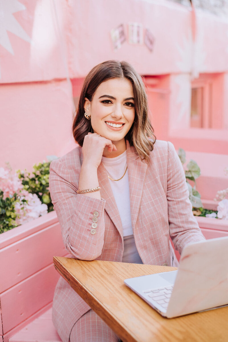Madison Kinnard, smiling and seated at an outdoor café in a pink suit, working on her laptop—representing her full-service digital support for women entrepreneurs.