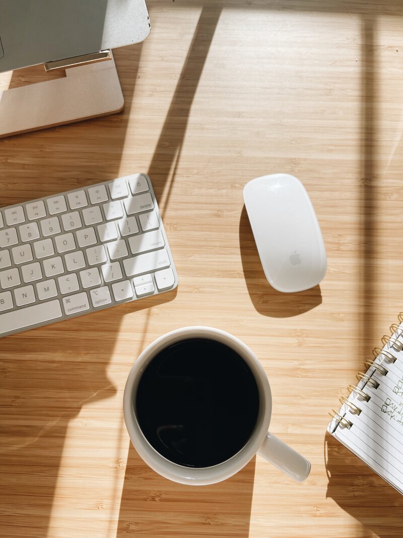 A desk surface with a keyboard, mouse, mug of coffee, and notebook