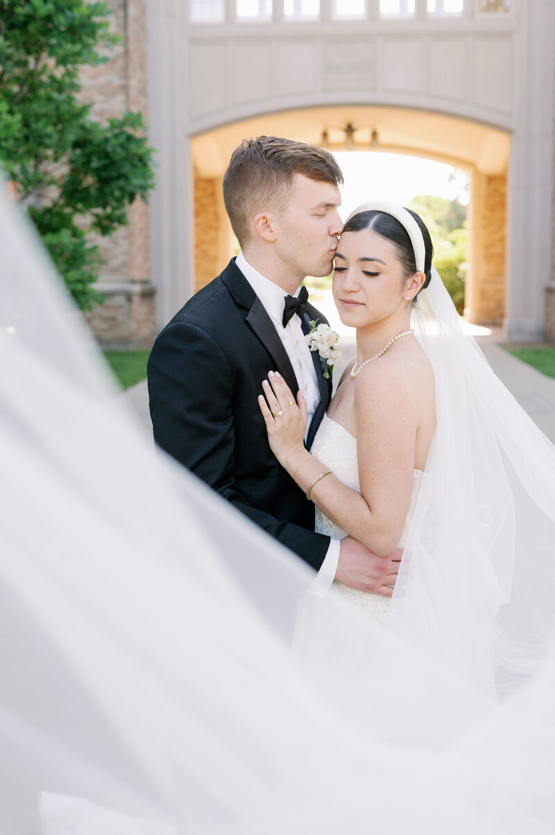 Groom kissing bride on the head while her veil flows in the wind