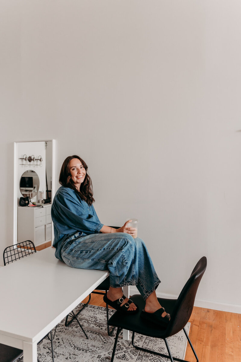 Delia, founder of Tidewater Studio, in a light studio space, sitting on a white desk with a denim top and jeans, iced latte in hand, smiling.